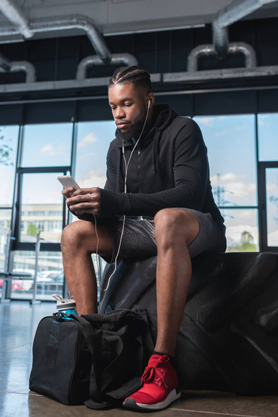 young african american man in earphones sitting in tyre and using smartphone in gym  - Φωτογραφία, εικόνα