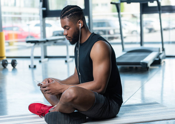 vista lateral del hombre afroamericano muscular en auriculares usando teléfono inteligente mientras está sentado en la esterilla de yoga en el gimnasio
 - Foto, Imagen
