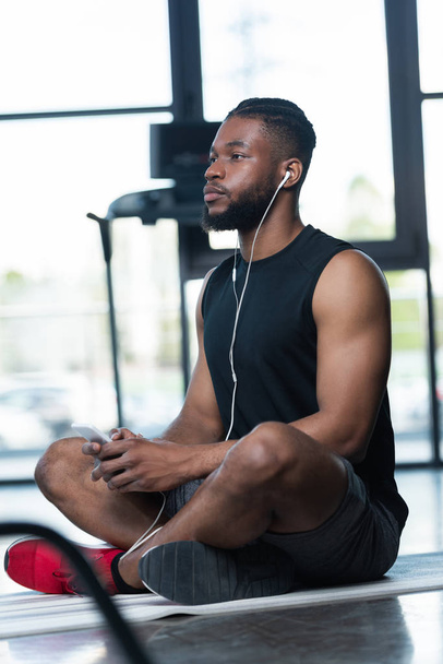deportista afroamericano pensativo en auriculares usando teléfono inteligente y mirando hacia otro lado mientras está sentado en la esterilla de yoga en el gimnasio
 - Foto, imagen
