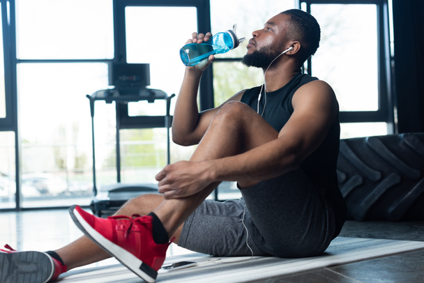 young african american sportsman in earphones sitting on yoga mat and drinking water in gym - Fotografie, Obrázek