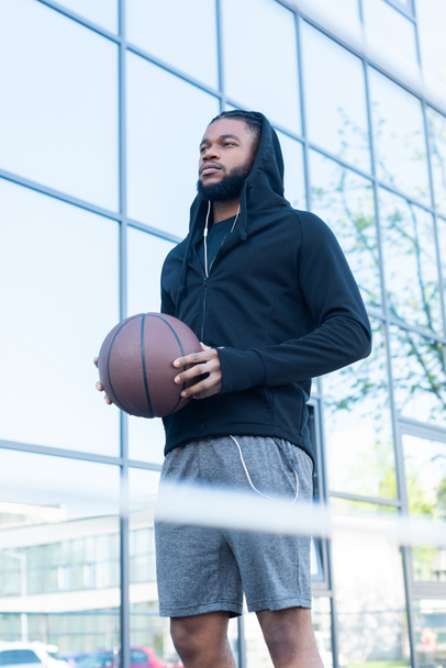 low angle view of young african american sportsman holding basketball ball and looking away on street - Fotoğraf, Görsel