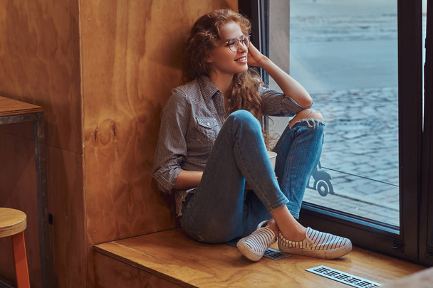 Smiling young redhead curly girl wearing casual clothes and glasses sitting on a window sill with a takeaway coffee. - Photo, Image