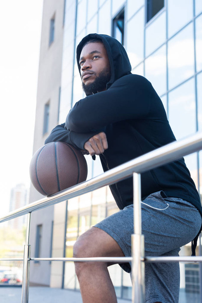 nadenkend Afro-Amerikaanse man leunend op basketbal bal en weg op zoek op straat - Foto, afbeelding