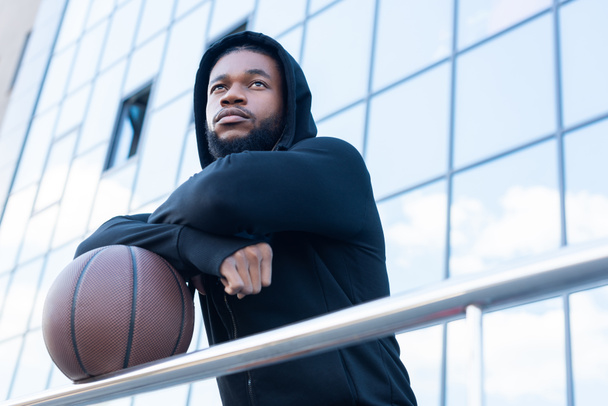 vista de ángulo bajo del pensativo deportista afroamericano mirando hacia otro lado mientras está de pie con pelota de baloncesto en la calle
 - Foto, Imagen