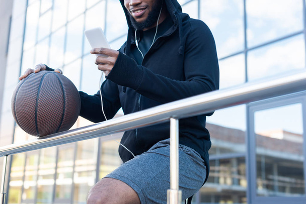 przycięte strzał uśmiechający się african american man w słuchawki za pomocą smartphone trzymając piłkę do koszykówki na ulicy - Zdjęcie, obraz