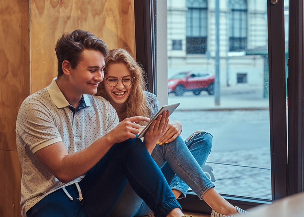 休憩中に大学の食堂で窓枠に座ってデジタル タブレットを使用して幸せな若い学生のカップル. - 写真・画像