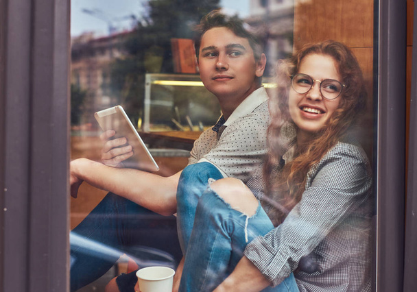 Jóvenes estudiantes felices tomando café y usando una tableta digital sentados en un alféizar de la ventana en un campus universitario durante un descanso
. - Foto, Imagen