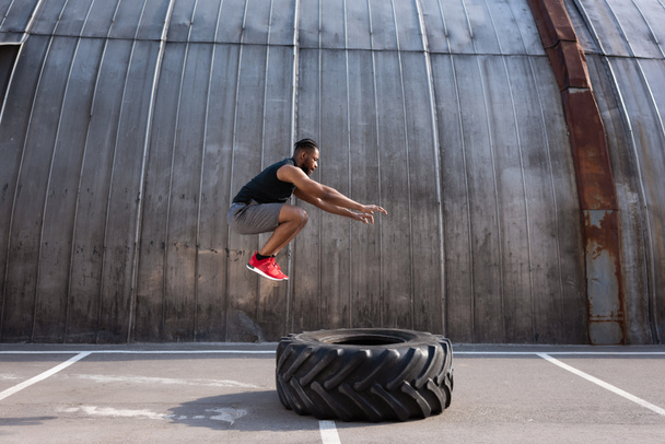 muscular african american sportsman jumping while training with tyre on street  - Foto, imagen