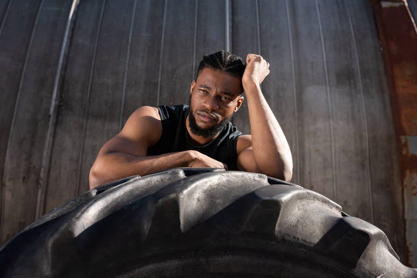 handsome young african american sportsman leaning at tyre and looking at camera - Foto, imagen