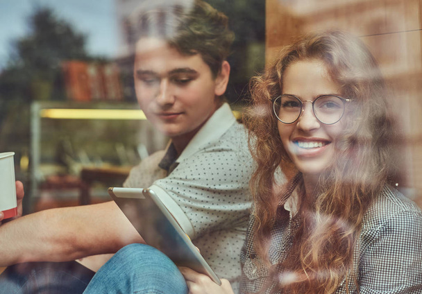 Счастливые молодые студенты пьют кофе и используют цифровые планшеты, сидя на подоконнике в университетском городке во время перерыва
. - Фото, изображение