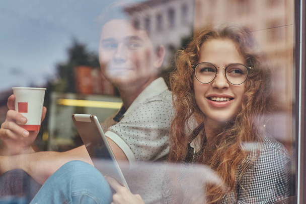 Jóvenes estudiantes felices tomando café y usando una tableta digital sentados en un alféizar de la ventana en un campus universitario durante un descanso
. - Foto, imagen