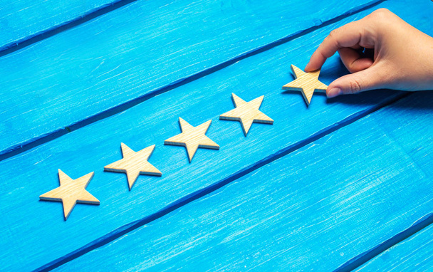 女性の手は、青色の背景に 5 番目の木製の星を置きます。評論家は、品質評価を設定します。5 つ星、最高品質マークです。ホテル、レストラン、カフェの評価レベルを上げる - 写真・画像