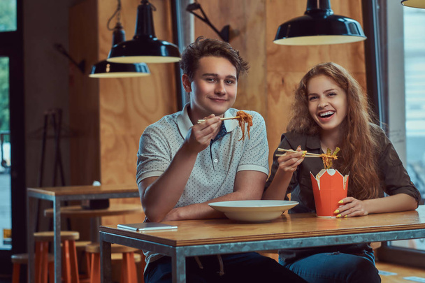 Heureux jeune couple portant des vêtements décontractés manger des nouilles épicées dans un restaurant asiatique
. - Photo, image