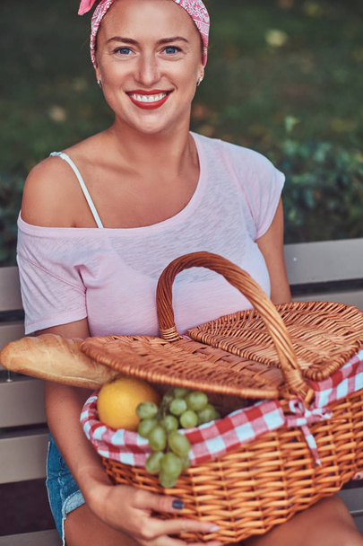 Happy beautiful redhead female wearing casual clothes holds a picnic basket while sitting on a bench in the park. - Фото, изображение