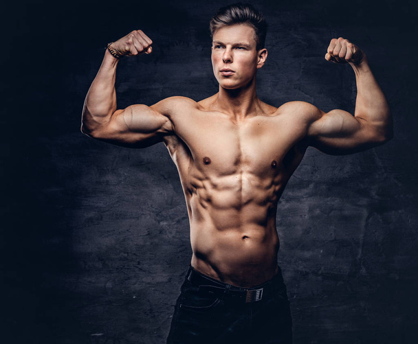 Сильный молодой мужчина без рубашки модель с красивым мускулистым телом показывает свой бицепс в студии. Изолированный на тёмном фоне
. - Фото, изображение