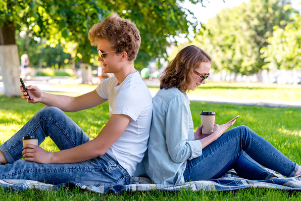 Students are a guy with a girl. In the summer in the park. He holds tea in his hands. He has a rest after school of institute. Looks at the smartphone. To correspond in social networks. - Foto, afbeelding