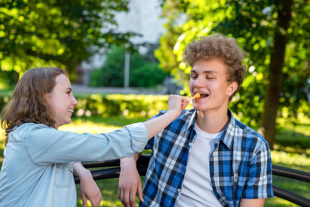 Young couple in love. Summer in park sit on a bench. A girl is feeding a guy with food. The guy smiles happily. The concept of a happy relationship. - Foto, Imagem