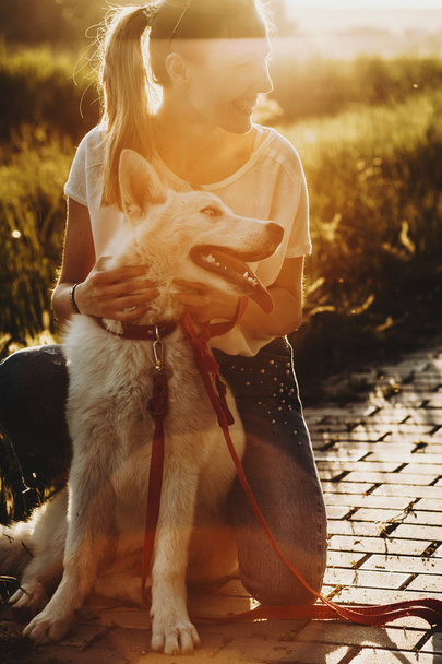 glückliche Frau in Sommerkleidung, die auf dem Gehweg auf Bunkern sitzt und einen weißen Hund umarmt, der wegschaut und bei Gegenlicht lacht - Foto, Bild