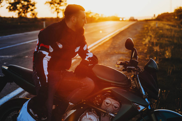 Widok boczny przystojny Brodaty mężczyzna w sprzęt ochronny, siedząc na stojący na siedzeniu drogowa motocykl z kasku w ręku niewyraźne tło podświetlane kraju trasy  - Zdjęcie, obraz
