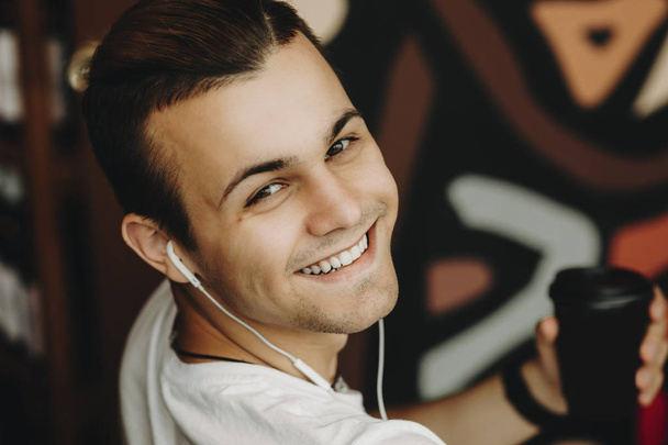 Upraw zbliżenie przystojny szczęśliwy młody mężczyzna w słuchawki trzymając kubek papierowy czarny toczenia przez ramię i patrząc na kamery na niewyraźne tło z kolorowym wzorem - Zdjęcie, obraz