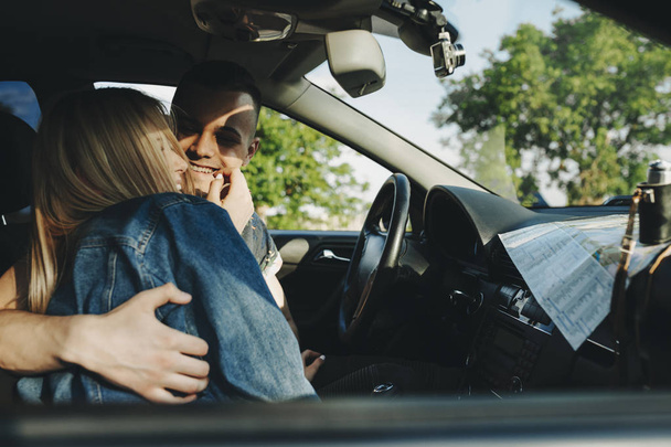 Вид збоку усміхненої блондинки, яка обіймає і тримає руку перед обличчям веселого молодого чоловіка, що сидить на кермі автомобіля з картою, що лежить на приладовій панелі
 - Фото, зображення