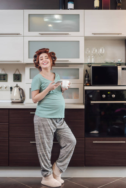 Wesoły, uśmiechnięty, w ciąży kobieta za pomocą smartfona, patrząc do aparatu trzymając kubek kawy lub herbaty w kuchni.  - Zdjęcie, obraz