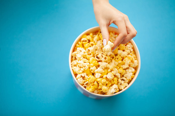 Élelmiszer. Nő kéz veszi Popcorn vödör. Pattogatott kukorica vödör a kék háttér. Mozi. - Fotó, kép