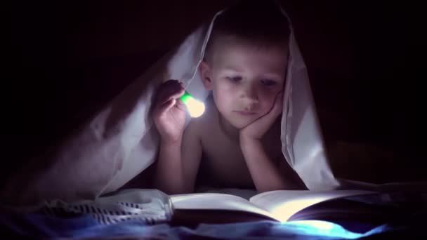 Uma criança lê um livro sob cobertores com uma lanterna à noite. menino com cabelo claro e olhos azuis
 - Filmagem, Vídeo