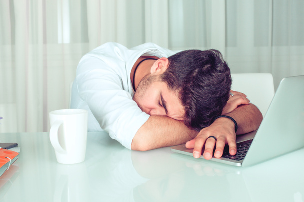 Liikemies nukkuu kannettavallaan toimistossaan kotona tietokoneella, herätyskello, paperit ja kirjat pöydällä
 - Valokuva, kuva