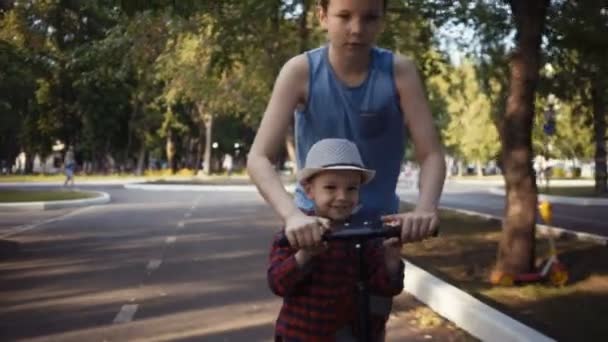 Два брата катаются на скутере в летнем парке
. - Кадры, видео