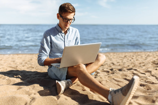 Young guy with glasses, working on his laptop on the beach, work on vacation, - Фото, зображення