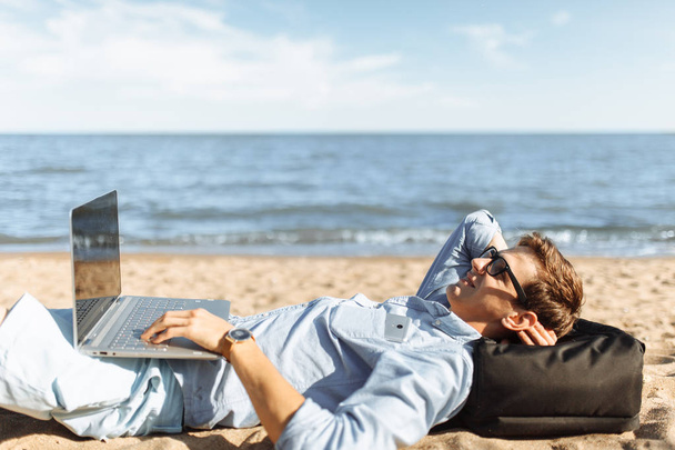 Young guy with glasses, lying on the sand, working on his laptop on the beach, against the sea, working on vacation, - Fotoğraf, Görsel