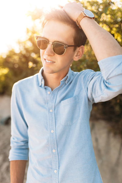 Beautiful stylish guy with glasses, posing on the beach, at sunset, hipster posing in stylish clothes, - 写真・画像
