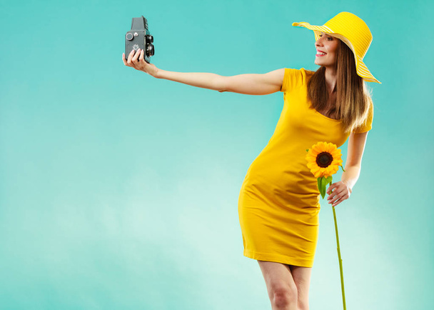 summer woman wearing yellow dress and hat with sunflower taking self picture with old vintage camera on vivid blue background - Photo, image