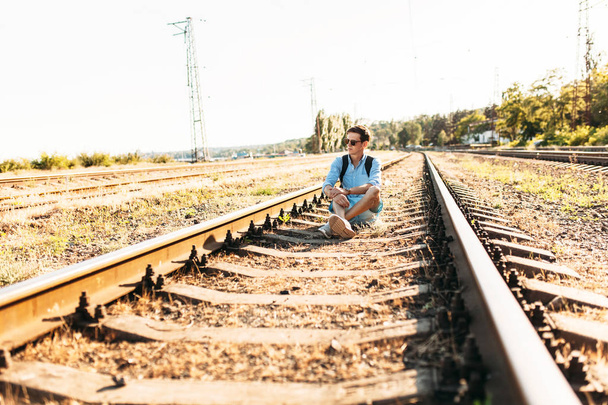 Beautiful stylish guy with glasses, posing sitting on the rails of the train at sunset, hipster posing in stylish clothes, - Fotografie, Obrázek