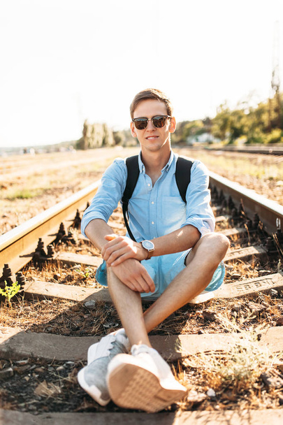Beautiful stylish guy with glasses, posing sitting on the rails of the train at sunset, hipster posing in stylish clothes, - Fotó, kép