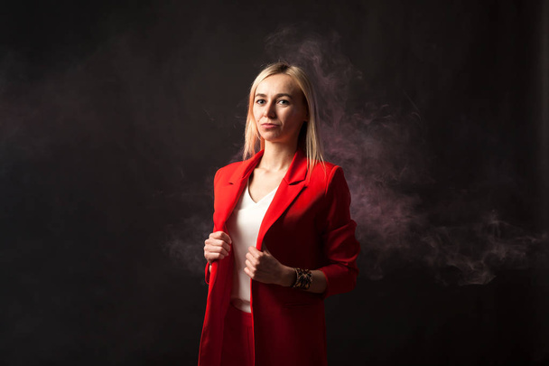 Young beautiful white blond girl in a bright red strict suit with a jacket and white blouse looks at the camera on a background of colored blue and pink smoke on a black isolated background - Photo, image