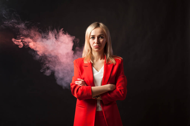 Young beautiful white blond girl in a bright red strict suit with a jacket and white blouse looks at the camera on a background of colored blue and pink smoke on a black isolated background - Photo, Image