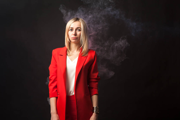 Young beautiful white blond girl in a bright red strict suit with a jacket and white blouse looks at the camera on a background of colored blue and pink smoke on a black isolated background - Photo, Image