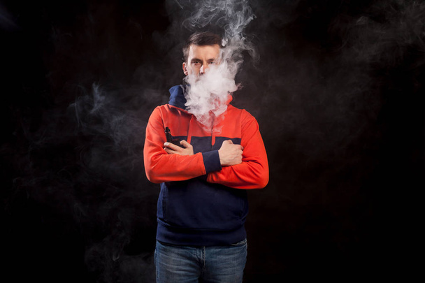 A young dark-haired man in a colored orange and blue hoody standing in close posture smokes a vape and exhales a white smoke on a black isolated background - Foto, Bild