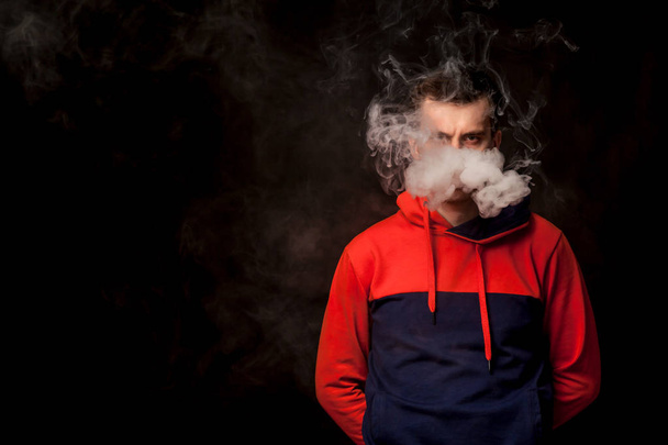 A young dark-haired man with sad eyes in a colored orange and blue hoody standing with arms behind smokes a vape and exhales a white smoke on a black isolated background - Фото, зображення