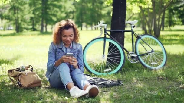 Happy African American girl is using smartphone touching screen and smiling while resting in park after riding bike. Modern bag and bicycle are visible. - 映像、動画