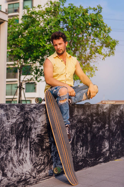 Young handsome man with beard sitting with longboard on the street. - Foto, afbeelding