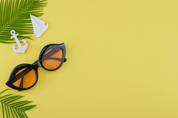 Солнечные очки украшают листья папоротника и крошечный парусник и якорь на желтом фоне с копировальным пространством
 - Фото, изображение
