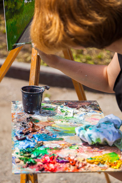Um artista de close-up pinta em uma tela uma paisagem urbana de um dique de verão com atracções de tintas a óleo
 - Foto, Imagem