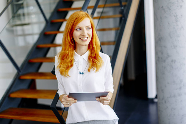 Όμορφη νεαρή κόκκινα μαλλιά γυναίκα χρησιμοποιώντας ψηφιακό tablet στο σύγχρονο γραφείο - Φωτογραφία, εικόνα