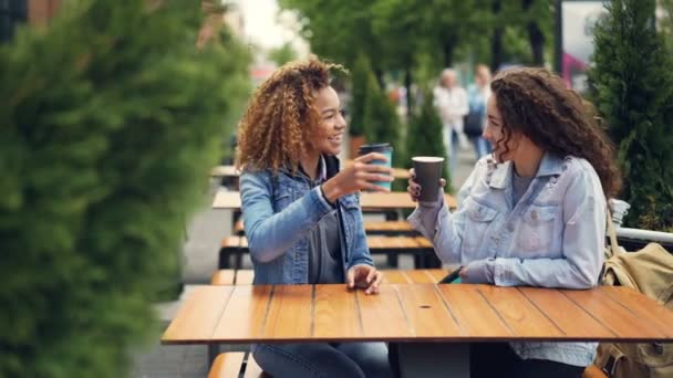 Hübsche junge Frauen rösten und klimpern Kaffeetassen, trinken Kaffee und unterhalten sich, während sie sich im Café im Stadtpark ausruhen. Menschen laufen im Hintergrund. - Filmmaterial, Video