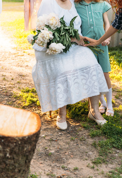 grootmoeder, een oudere vrouw in wit, vintage kleding zitten en knuffelen haar kleindochter, een meisje in een groene jurk, en die in haar andere hand een boeket van bloemen, witte pioenrozen, in de tuin. vergadering, gefeliciteerd en een gift.a vrouwelijke han - Foto, afbeelding