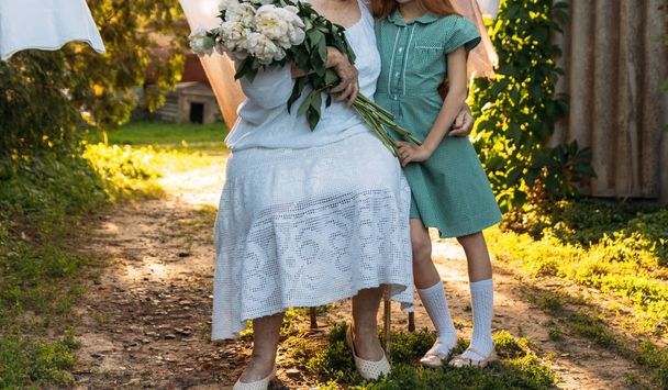 conversation between grandmother and granddaughter. grandmother, an elderly woman in white, vintage clothes sitting and hugging her granddaughter, a girl in a green dress, and holding in her other hand a bouquet of flowers, white peonies, in the gard - Photo, Image