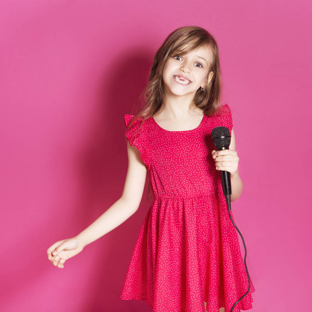 Little 8 years old girl make some emotional gesture with her hands on a pink neutral background. She has long brunette hair and wear red summer dress. Funny expression on her face - Fotografie, Obrázek